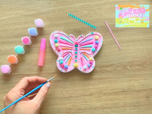 A persons hands can be seen with a paintbrush painting a plaster of Paris butterfly. Around this is Gem stickers, glitter glue and 6 paints.