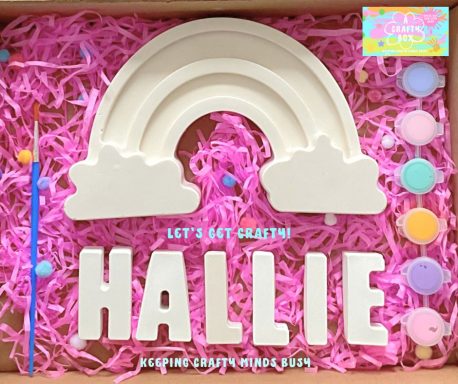 Large rainbow mould on top of pink tissue paper. Six paints in pastel colours to the right, under the rainbow it spells the name HALLIE. 