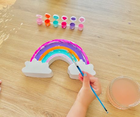 Child holding a paintbrush painting a large plaster of Paris rainbow in pink, purple, orange, teal and baby pink.