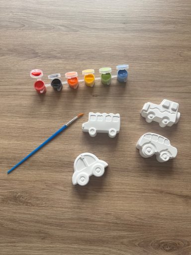 Four plaster of Paris cars on a wooden table. A paintbrush is next to this with six opened paints in various colours.